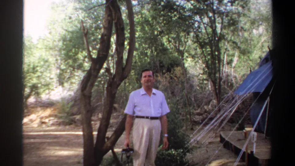 The man pauses near the blue tent in a shaded Khartoum campsite, framed by trees and tent lines.