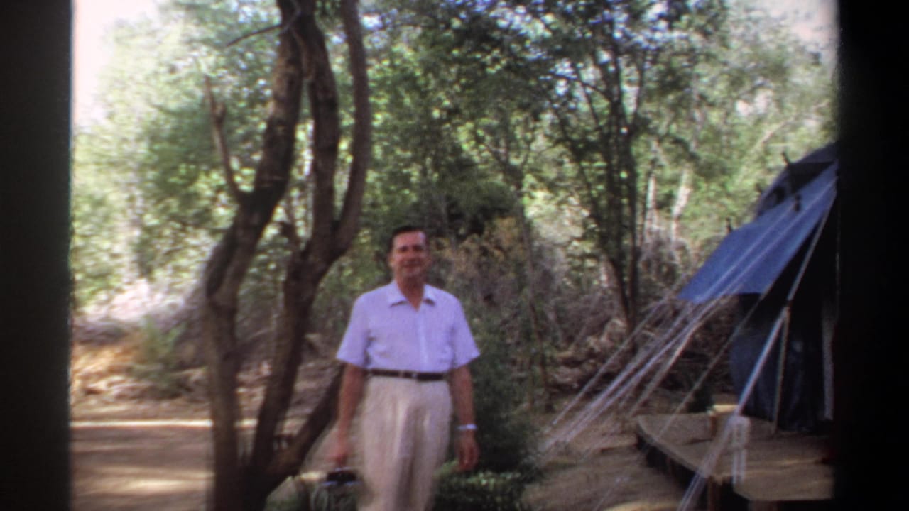Man in light clothing standing near a large blue tent in a wooded campsite.