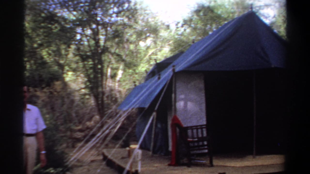 1969 Khartoum campsite view with a blue tent, guy ropes, trees, and a partially visible adult man.