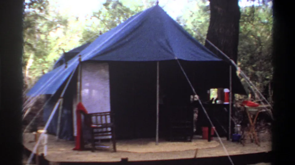 1969 Khartoum campsite frame showing a large blue tent, platform, camp chair, table, and supplies under trees.