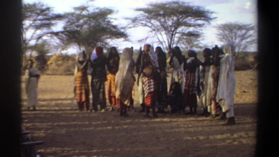 1969 Khartoum home-movie frame showing a large outdoor group in patterned wraps and head coverings under sparse trees.