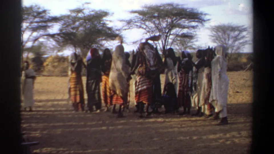 1969 Khartoum frame showing people gathered in a loose circle, some raising arms, on dry ground near trees.