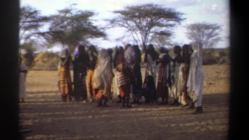 1969 Khartoum home-movie frame with a group in patterned clothing and head coverings gathered beneath trees.