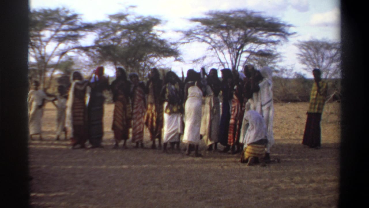 1969 Khartoum home-movie frame showing women in patterned and white garments gathered outdoors beneath sparse trees.