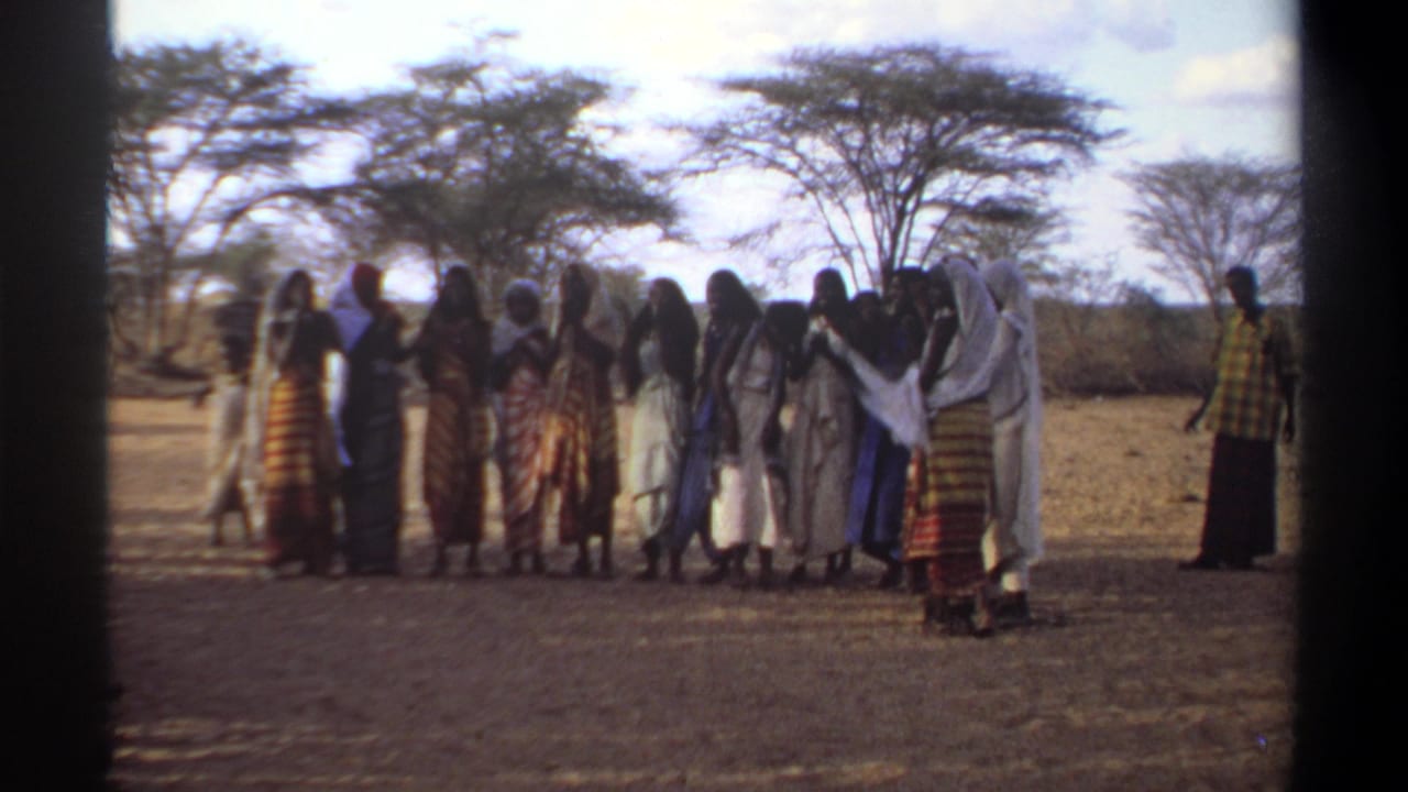 In the Khartoum 1969 home-movie frame, the clapping group remains lined up as a man in a checkered shirt observes nearby.