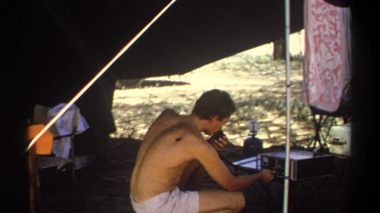 Shirtless man crouches under a tent beside a portable radio unit and microphone.
