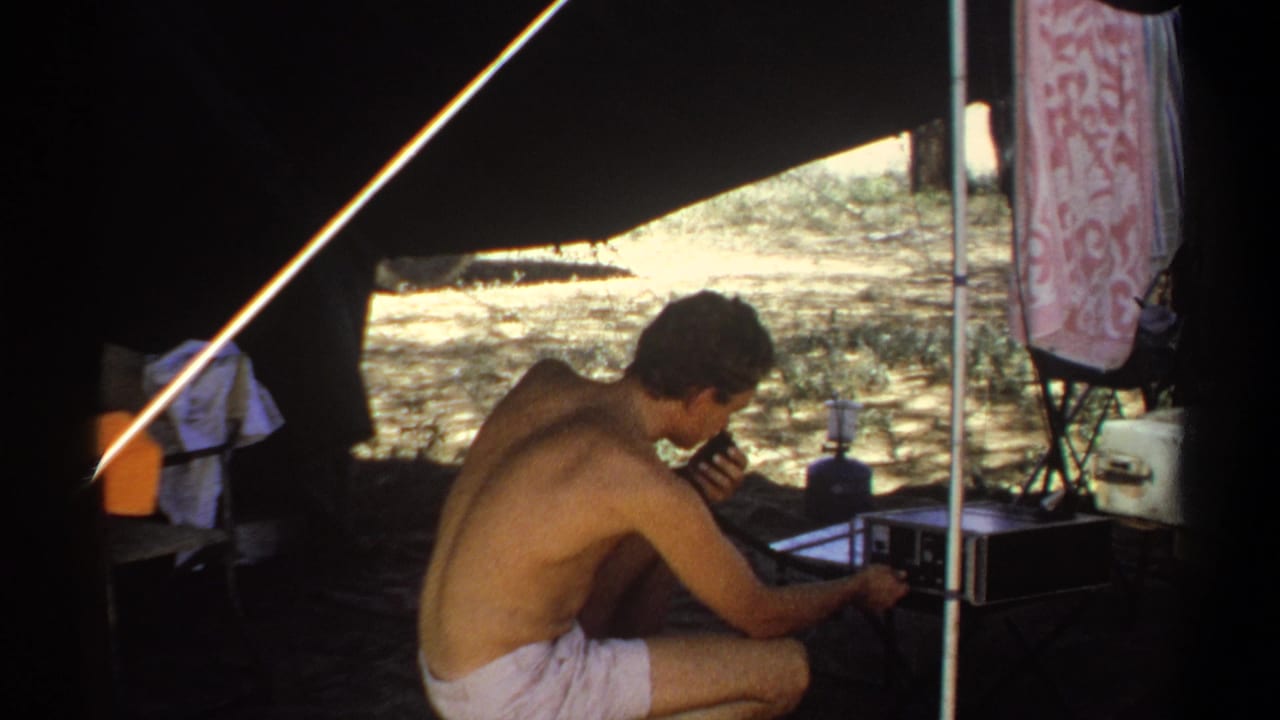 The man remains crouched under the tent in Khartoum, holding a microphone close while adjusting or monitoring a portable radio device.