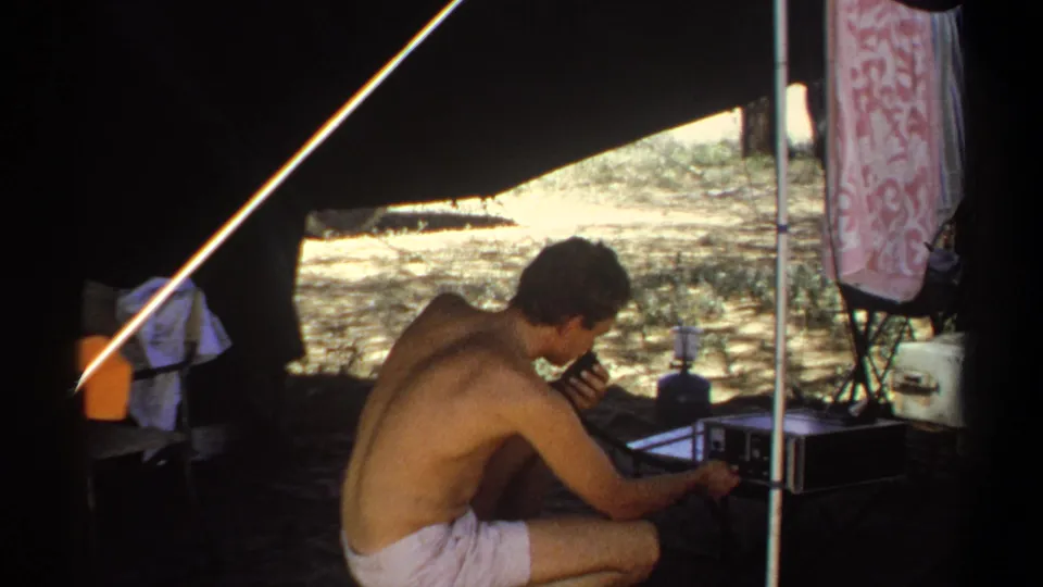 The man remains crouched under the tent in Khartoum, holding a microphone close while adjusting or monitoring a portable radio device.