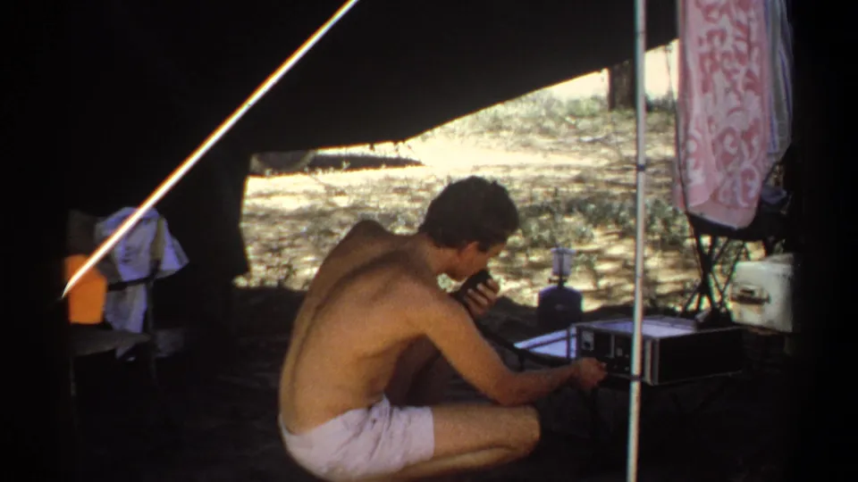 A shirtless man crouches low beside a portable radio set, with tent poles, cloth, and camping gear visible around him.
