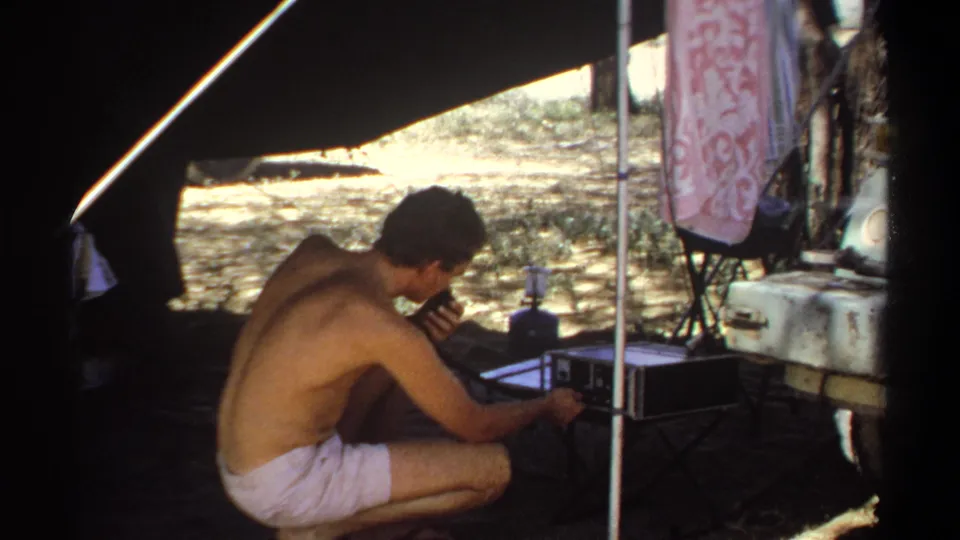The final frame shows the man still crouched at the radio unit beneath the tent, with a cooler and sunlit outdoor ground behind him.