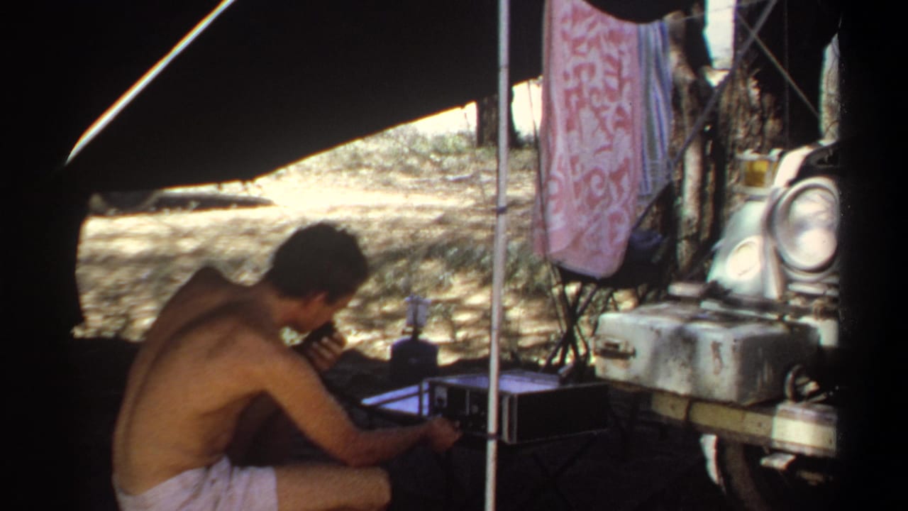 In 1969 Khartoum home-movie footage, a shirtless man sits under shade near portable equipment, a lantern, a hanging towel, and a parked vehicle.