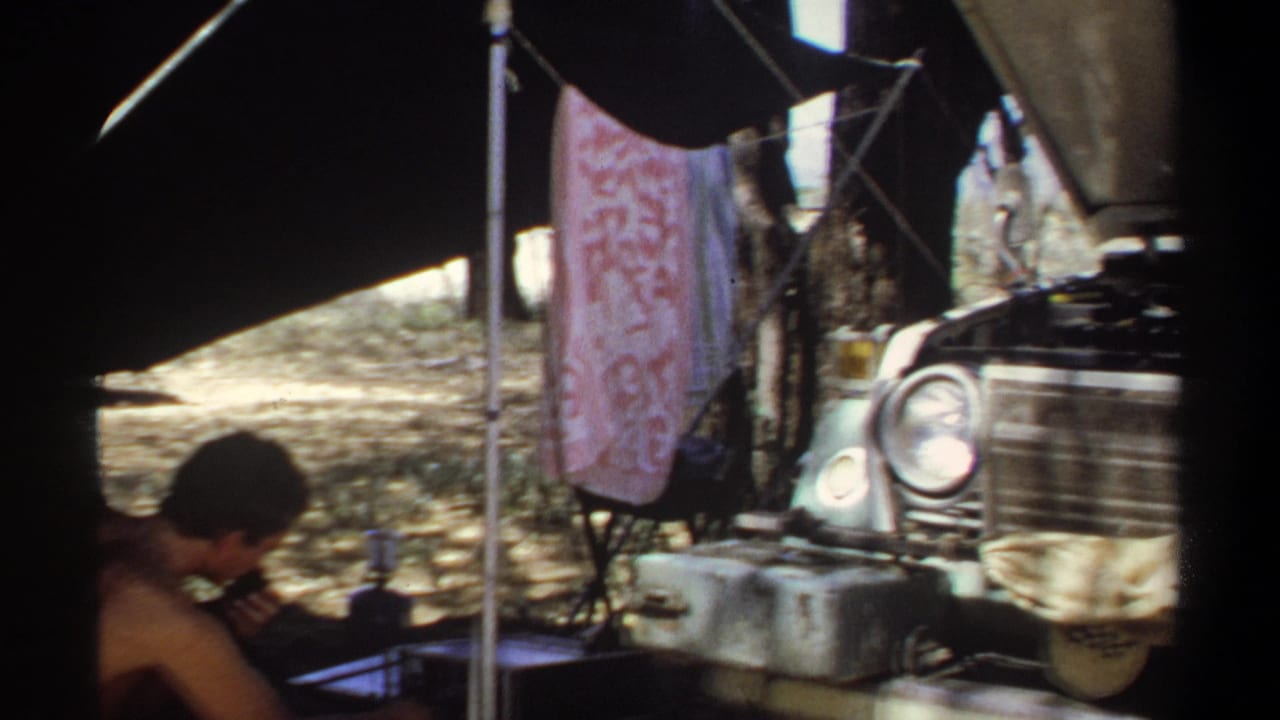 A man in the Khartoum camp sits near a radio or device while a towel hangs behind him and the front of an open-hood vehicle fills the side of frame.
