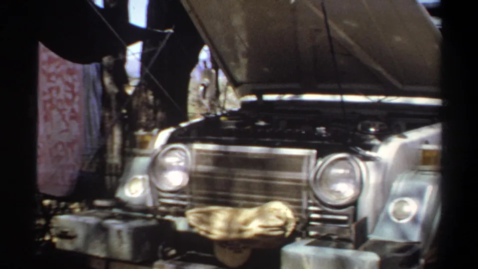 The 1969 Sudan camp footage centers on the front of an older vehicle with its hood open, with a patterned towel, camp gear, and shade structure nearby.