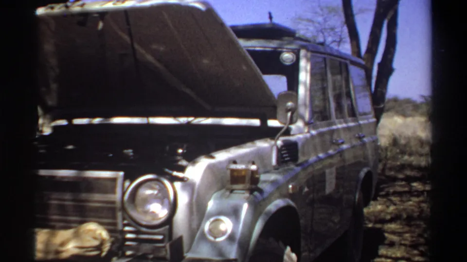 An older off-road vehicle stands with its hood raised in dry terrain near Khartoum, framed by trees and the dark edge of the 8mm gate.