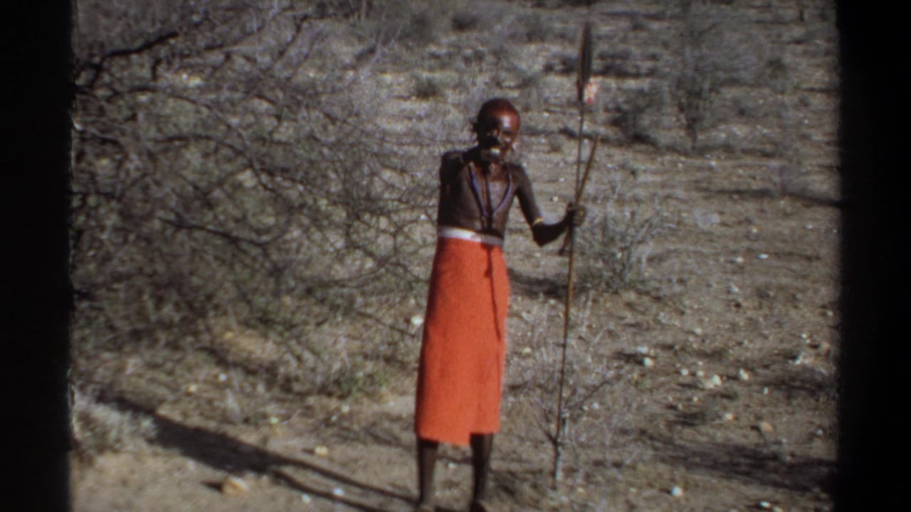 Man in an orange wrap stands in dry brush holding a spear.