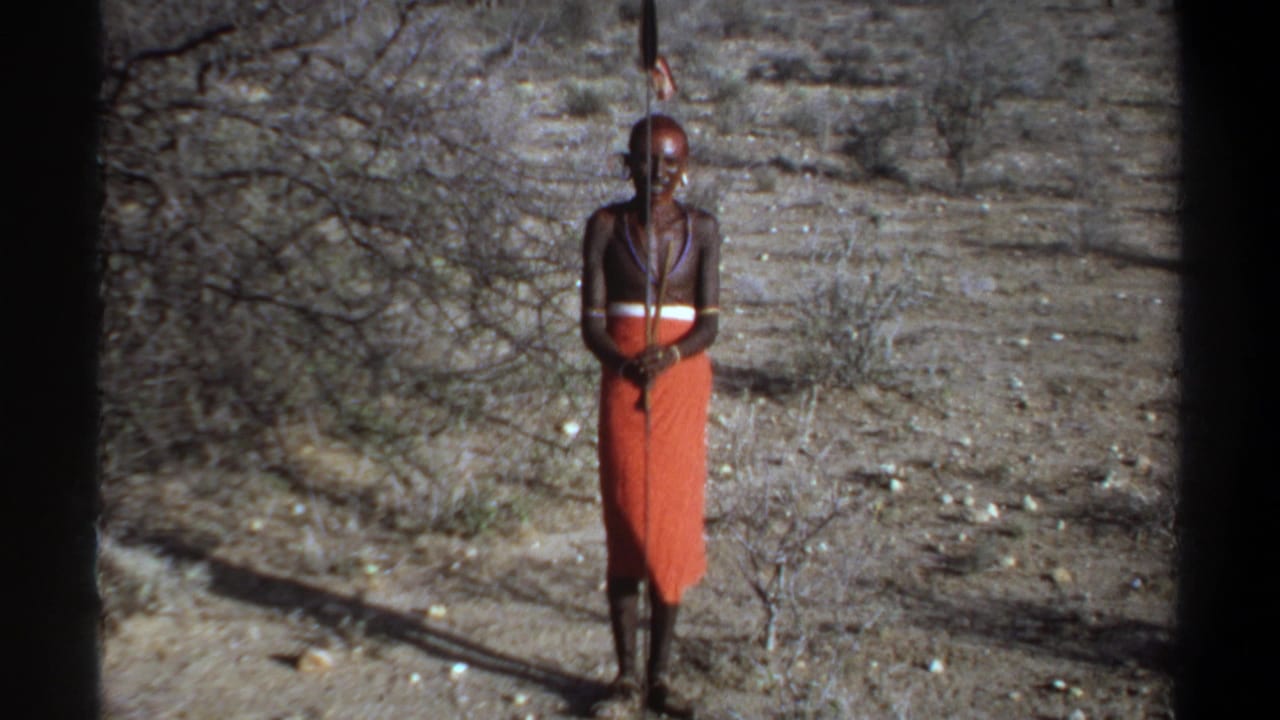 1969 Khartoum home-movie frame showing a man in an orange wrap garment, bead necklaces, and a long staff in dry brushland.