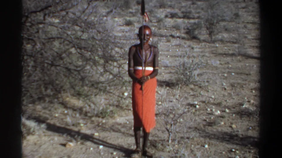 1969 Khartoum home-movie frame showing a man in an orange wrap garment, bead necklaces, and a long staff in dry brushland.