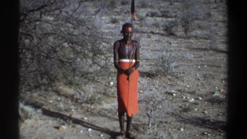 1969 Khartoum frame showing the man still facing the camera, staff vertical, with orange clothing and sparse brush behind him.