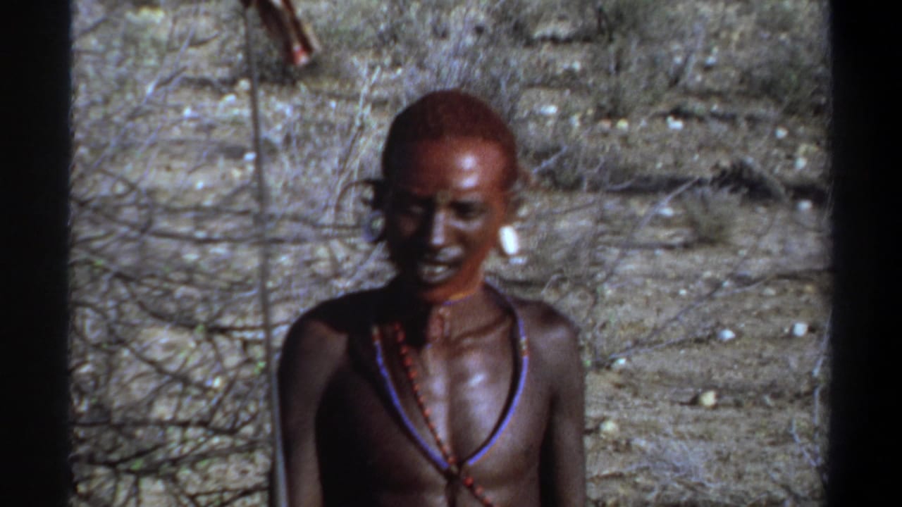 1969 Khartoum 8mm frame of an adorned man facing the camera, with white ear plugs, beads, bare shoulders, and scrubby dry ground behind him.