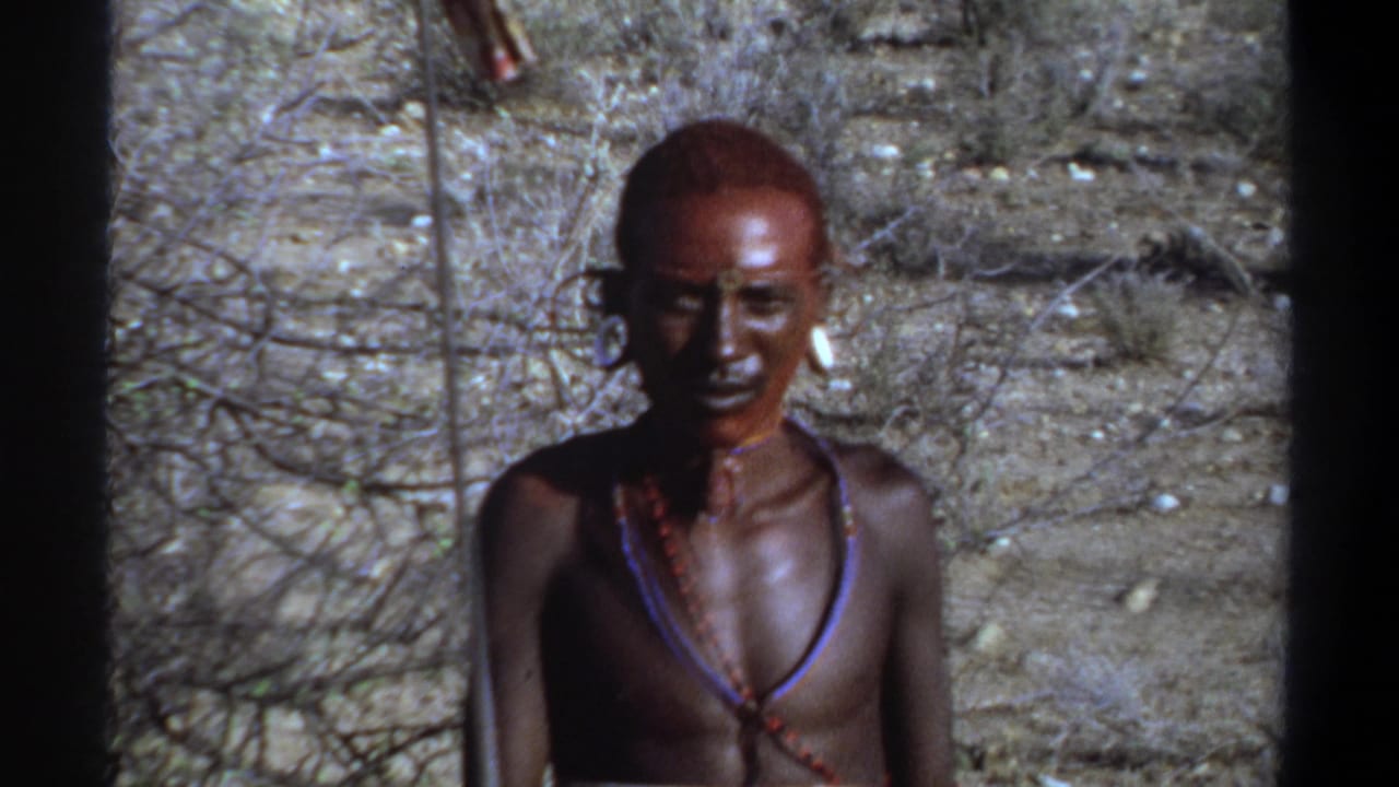 1969 Sudan frame showing a man in face paint and necklaces standing nearly still in bright daylight against sparse brush and dry earth.