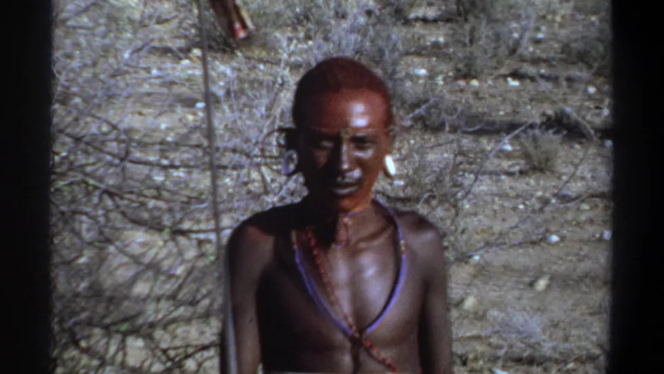1969 Sudan frame showing a man in face paint and necklaces standing nearly still in bright daylight against sparse brush and dry earth.