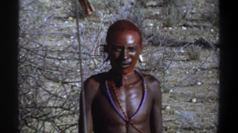 Khartoum 1969 8mm frame of a man with a shaved head, red face paint, white ear plugs, and beadwork, framed by dark film edges.