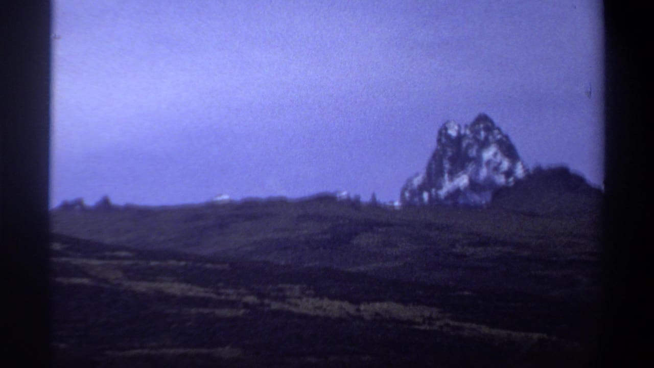 1969 8mm frame showing a distant snow-streaked mountain rising beyond dark rolling hills under a blue-purple sky.