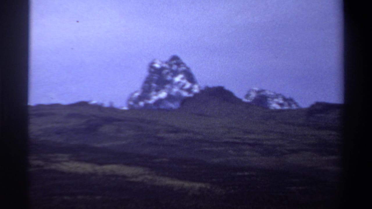 Snow-streaked peak and lower ridge seen beyond shadowed hills.