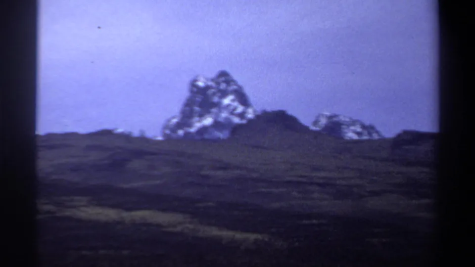 1969 8mm frame with a snow-streaked mountain and lower ridge beyond shadowed hills, tinted blue-purple by the film stock.
