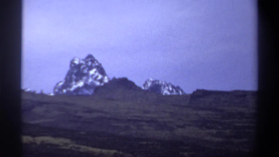 1969 8mm frame showing a jagged mountain with snow patches behind a dark hillside and muted grassy foreground.