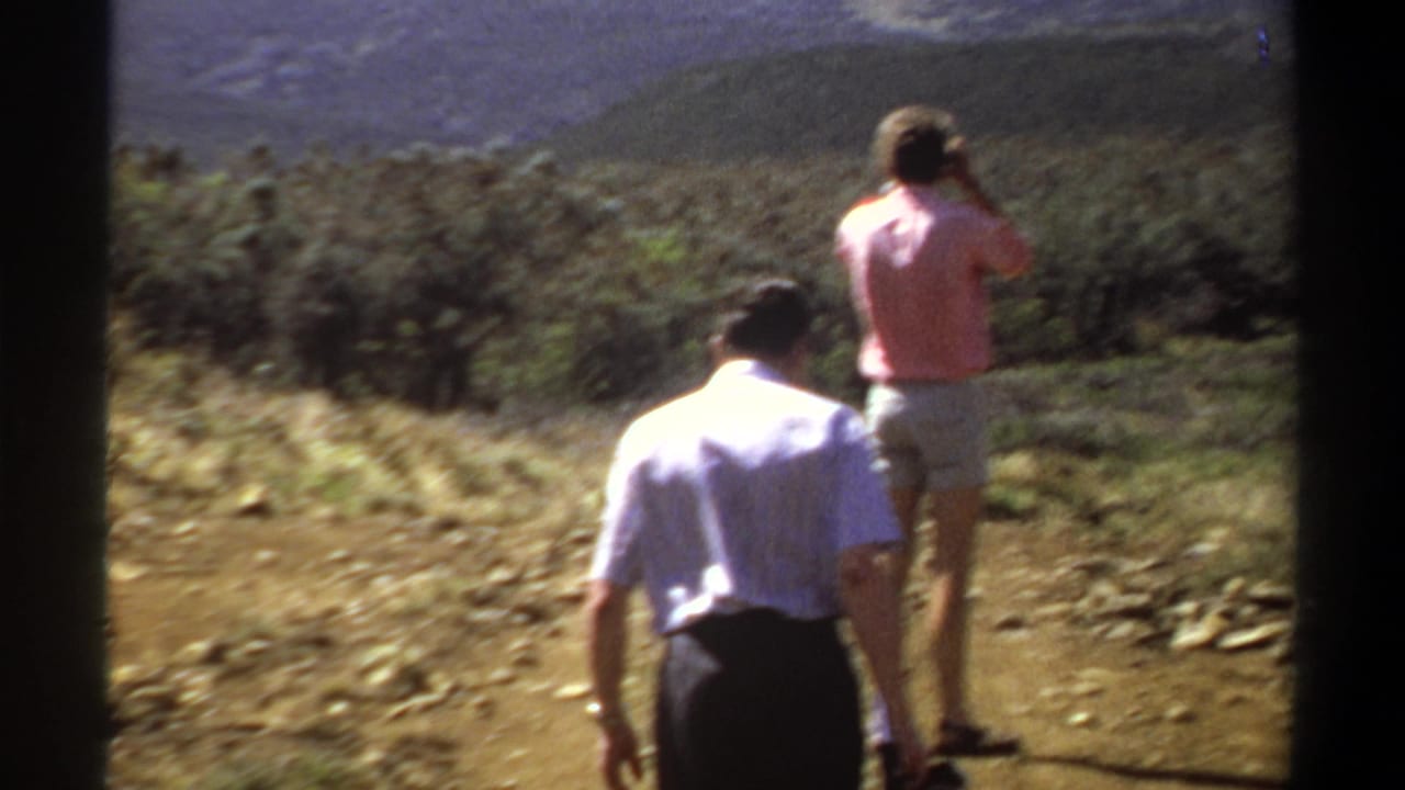 Two adult men on a Khartoum-area hillside in 1969, one in a pink shirt lifting a camera toward the distant mountain view.