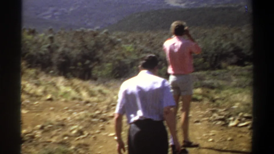 Two adult men on a Khartoum-area hillside in 1969, one in a pink shirt lifting a camera toward the distant mountain view.