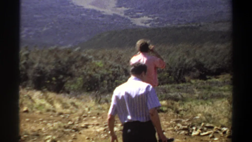 1969 home-movie frame from Sudan showing one man taking a landscape photo and another walking on a dry, rocky hillside.
