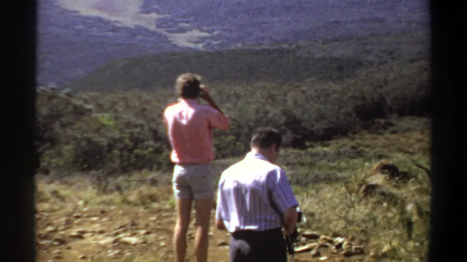 Two men stand on a rugged Khartoum-area hillside in 1969, with one holding a camera and dry brush leading toward hazy hills.