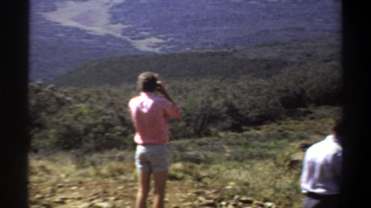 A woman in a pink shirt looks across a sunlit Sudan landscape while a second adult is partly cropped at right.