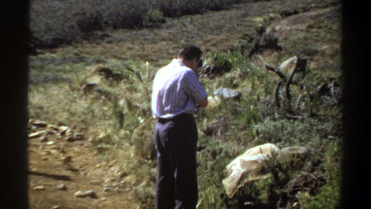 In 1969 Sudan home-movie footage, a man in a light shirt stands among rocks and grasses while photographing the countryside.