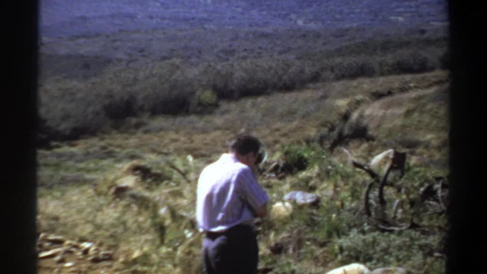 The 8mm frame shows a man in a pale shirt photographing from a dry, rocky grassland area in Sudan.