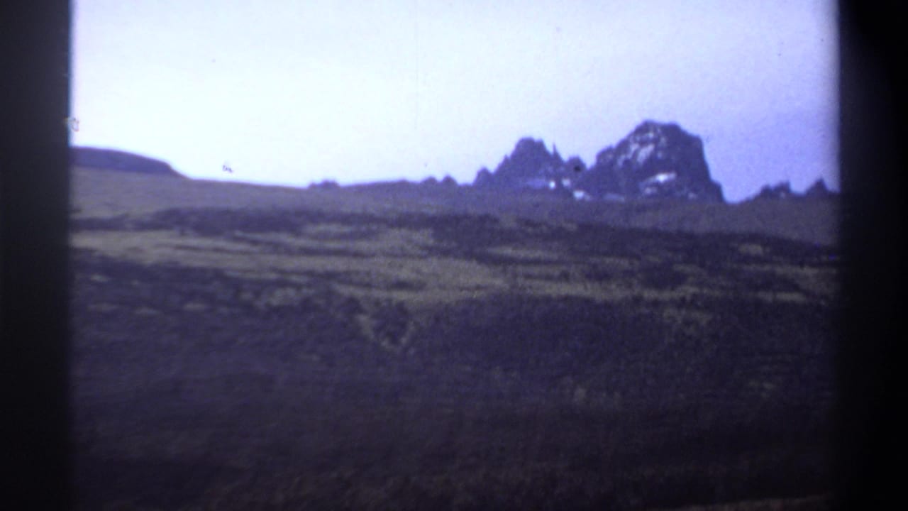 Distant rocky mountains beyond dark grassland under a pale sky.