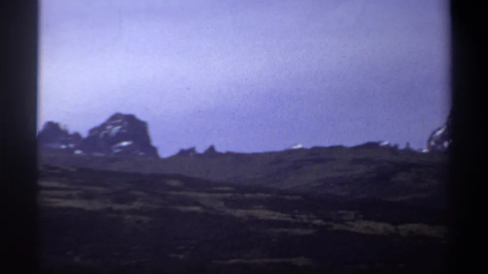 1969 8mm landscape frame with a grassy slope, low mountain ridge, and pale blue sky.