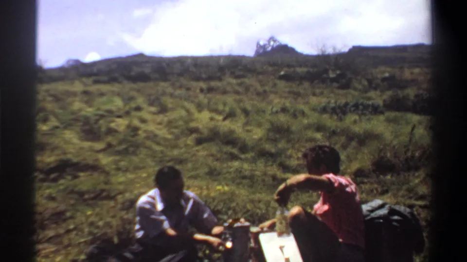 Two men in 1969 Khartoum, Sudan footage sit on grass near bottles and a box, with the hillside filling the background.