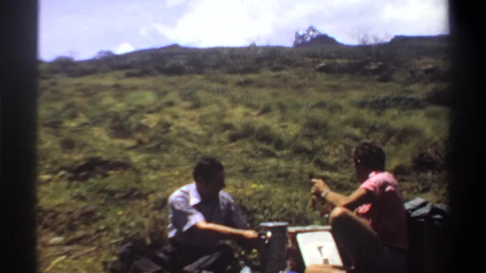 In the 1969 Sudan home-movie scene, one man sits near the pale box while the man in pink holds a bottle beside him.