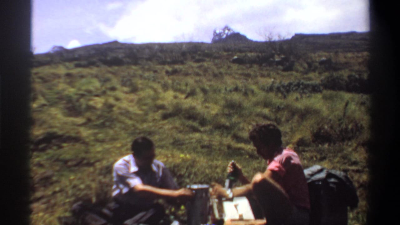 1969 Sudan frame showing a man in a light shirt and a woman in pink seated in grass during a hiking break.