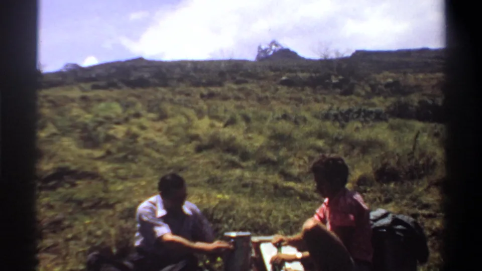 1969 Sudan home-movie frame of two adults seated close to hiking gear and drink containers on a sunlit grassy slope.