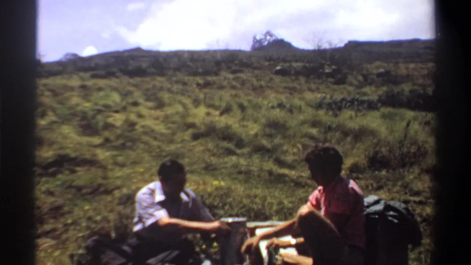 1969 Sudan frame of two hikers seated in profile near drinks and a backpack, with scrub-covered hills behind them.