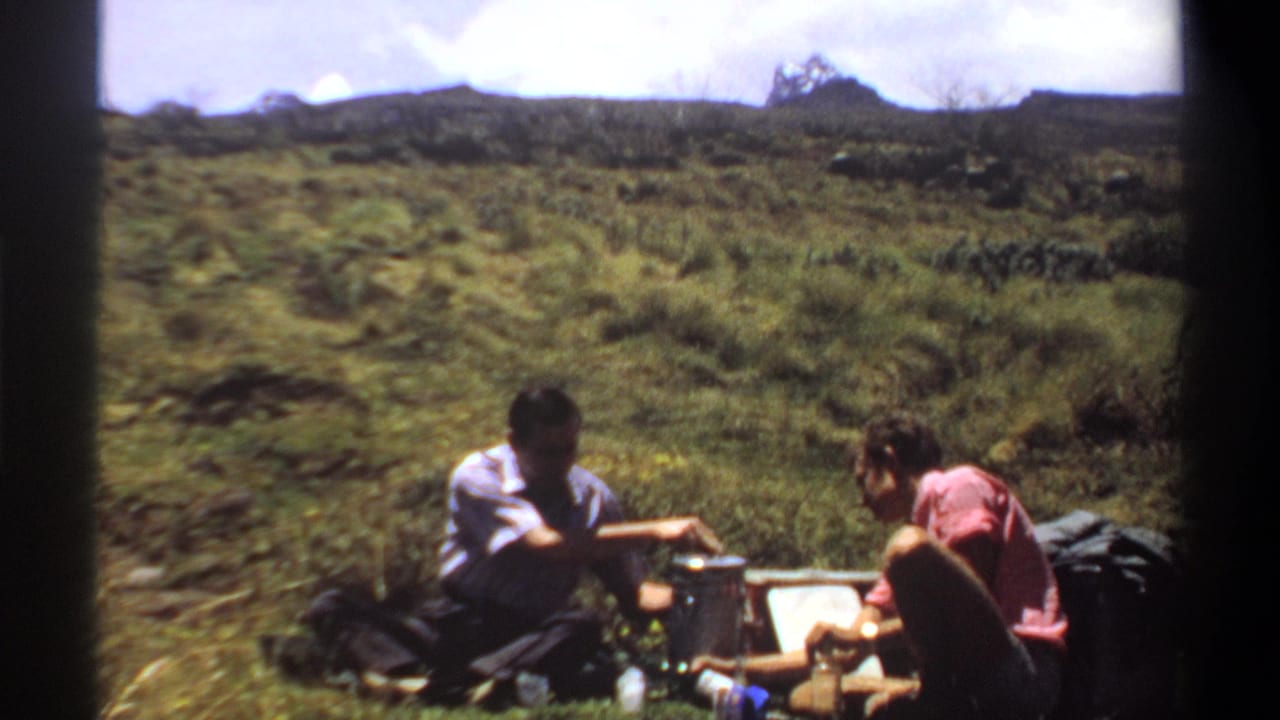 1969 Khartoum 8mm frame of two adults seated outdoors beside containers, bags, and a possible portable cooking setup.
