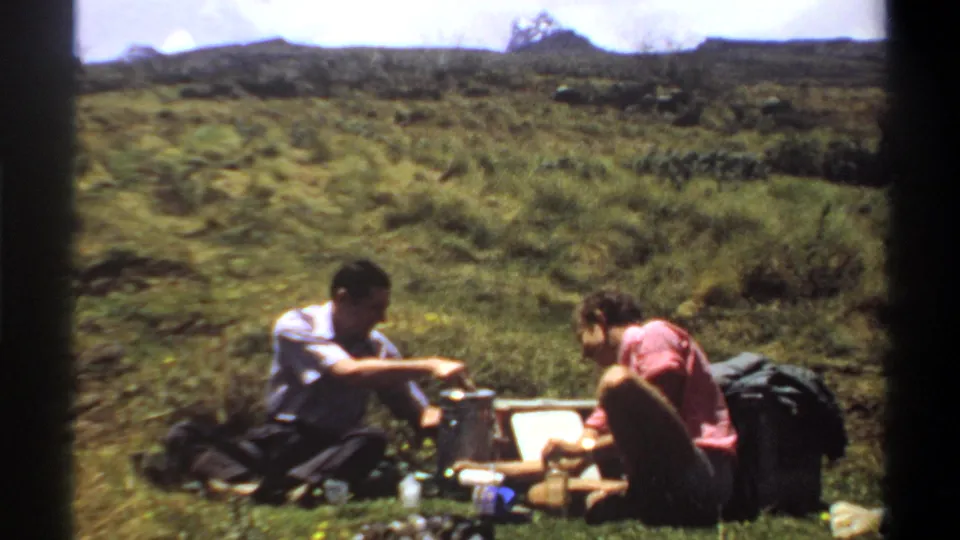 1969 Khartoum picnic frame with two adults seated low in the grass, surrounded by bags and small containers under a bright cloudy sky.