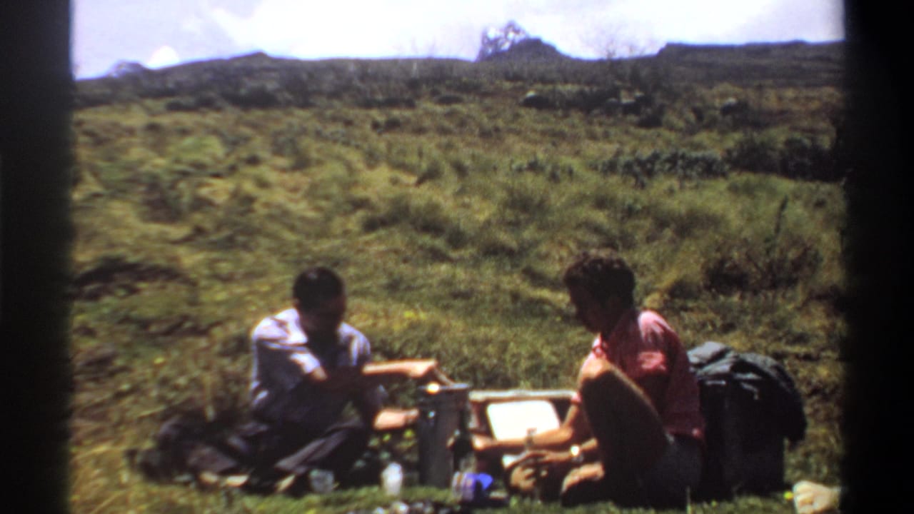 Two adult men in casual clothes sit beside picnic gear on grass in 1969 Khartoum-area home-movie footage.