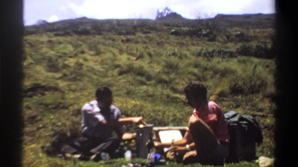 Two adult men in casual clothes sit beside picnic gear on grass in 1969 Khartoum-area home-movie footage.