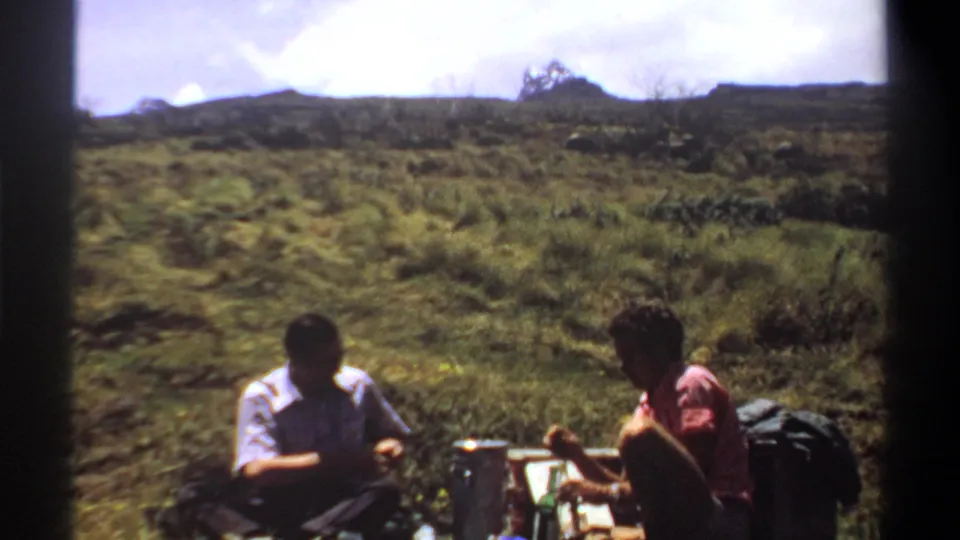 A soft 8mm frame shows two adult men seated with picnic supplies on grass near Khartoum, Sudan, in 1969.