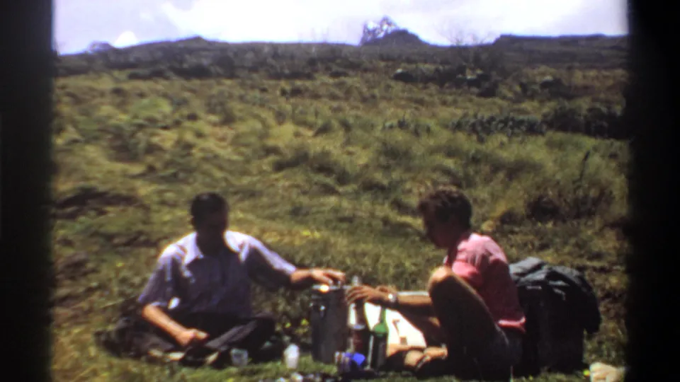 Two adult men reach between picnic containers and bottles on grass, with a wide Khartoum-area landscape behind them.